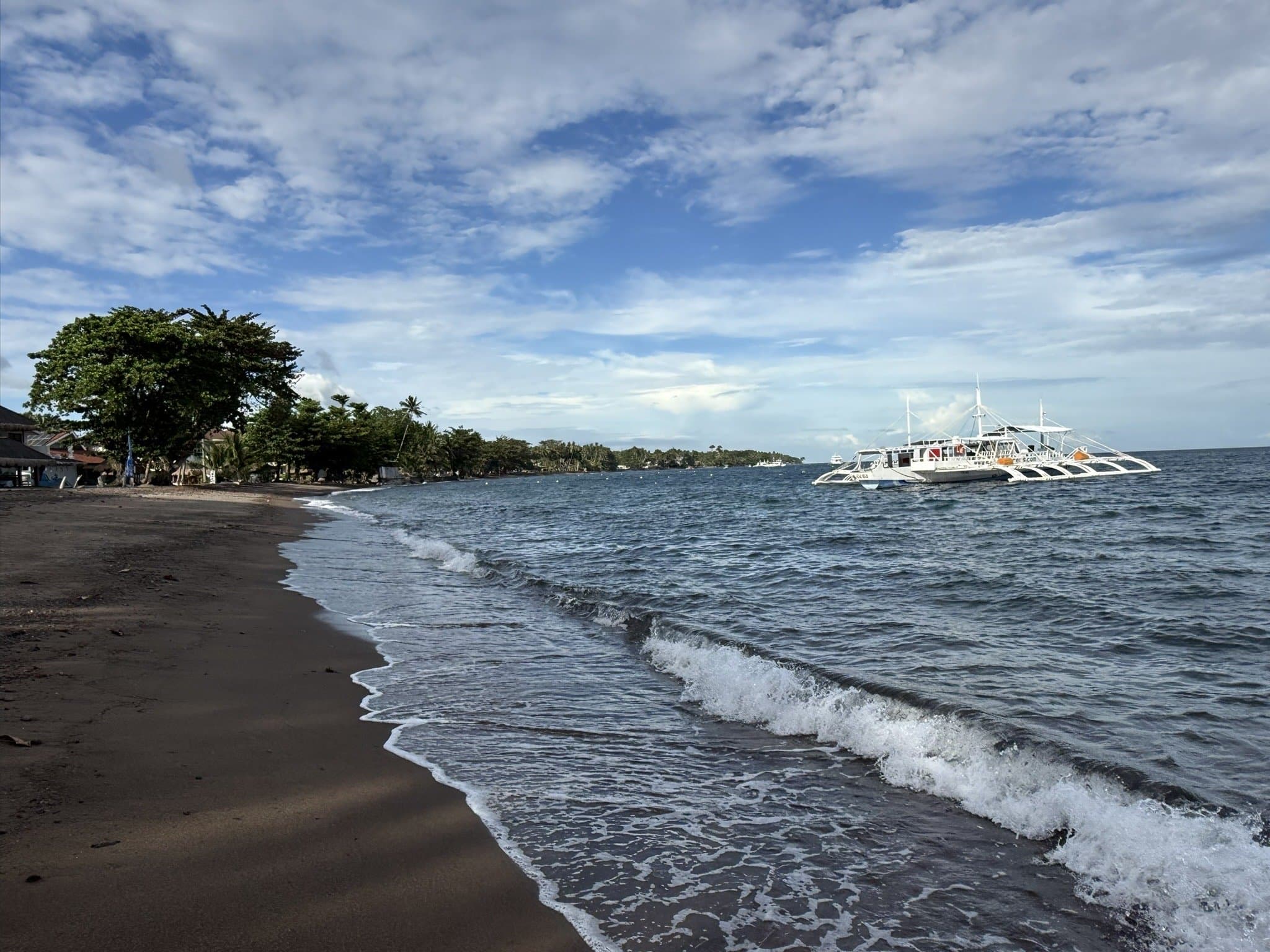 Dauin's black volcanic sand beach — a banca moored offshore, the dive sites directly below the surface in front of the resort