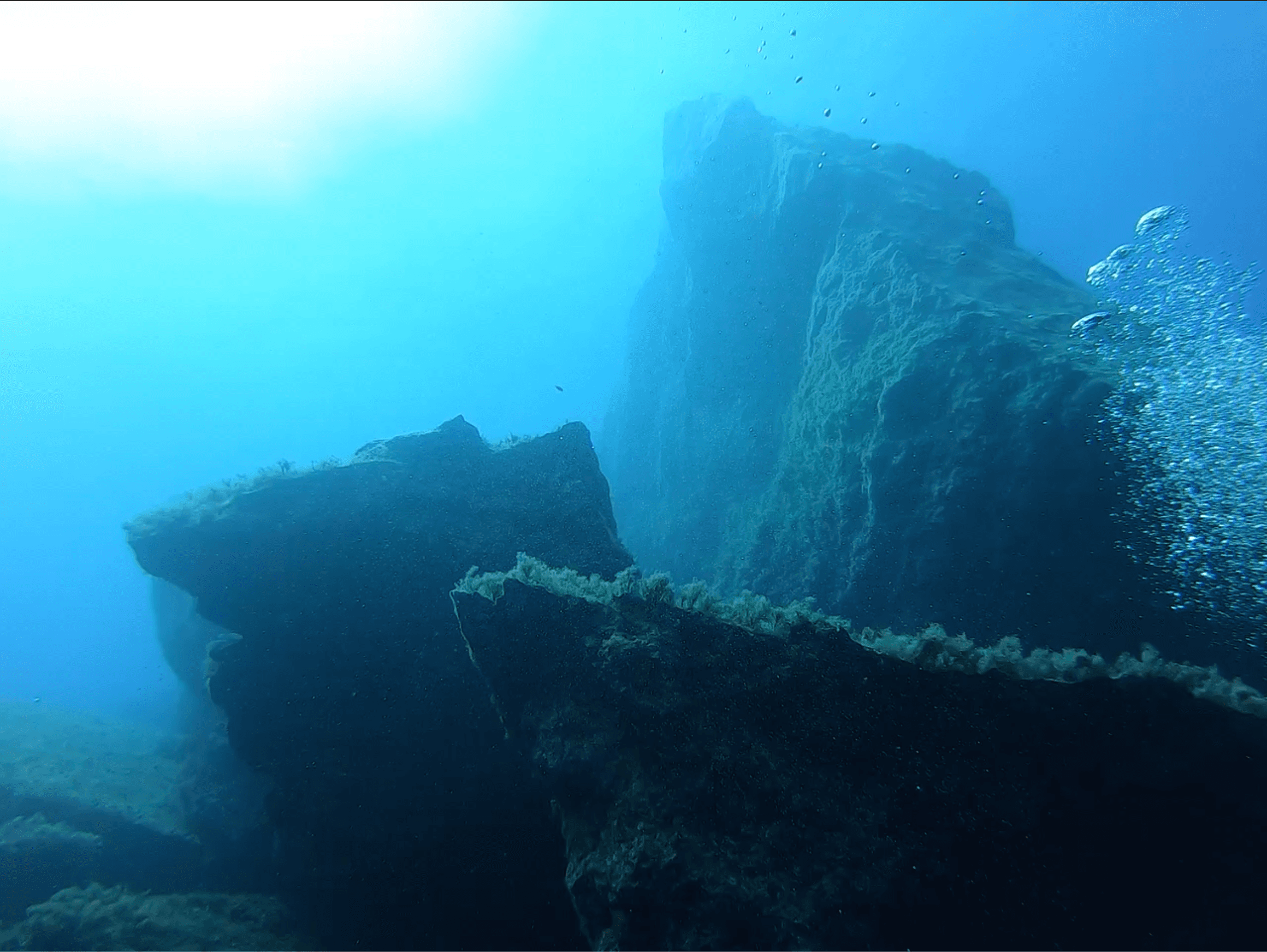 The Blue Hole at depth — rocky structure at 25 metres with small fish schooling in the haze, sunlight filtering from the chimney above