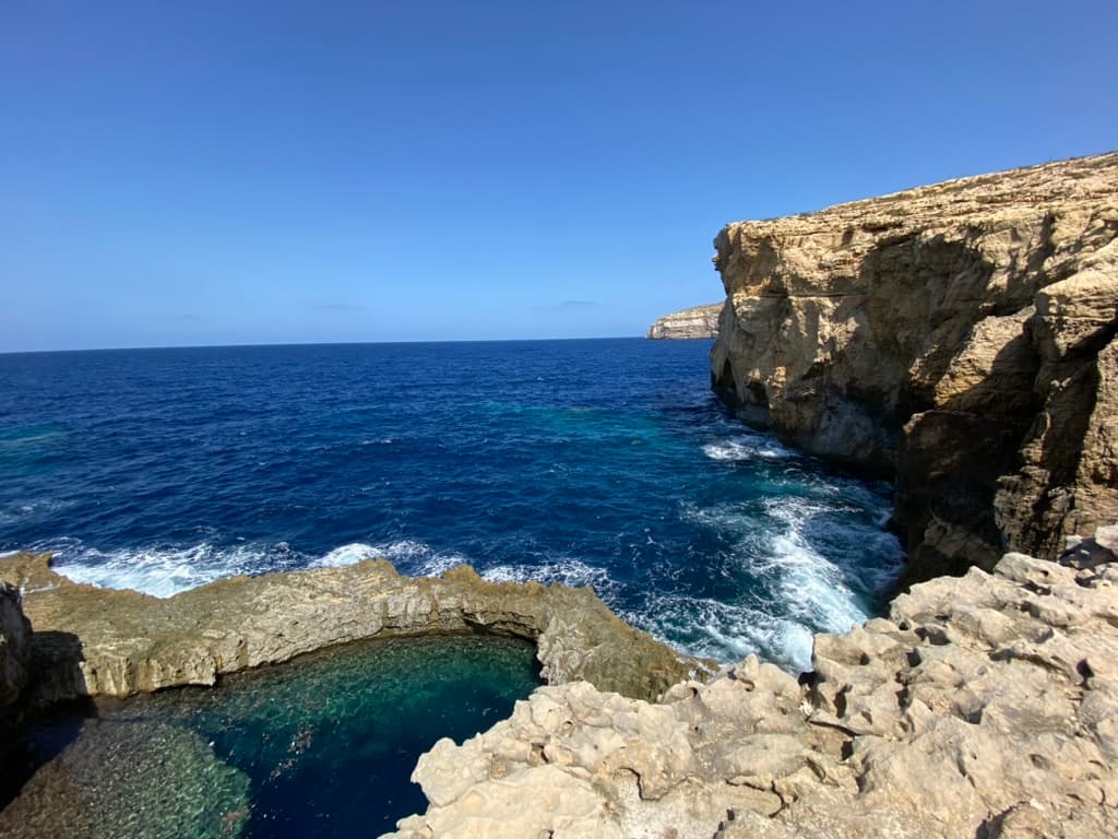 Dwejra Bay from the cliff top — the Blue Hole pool visible in the foreground limestone, the Mediterranean open to the horizon