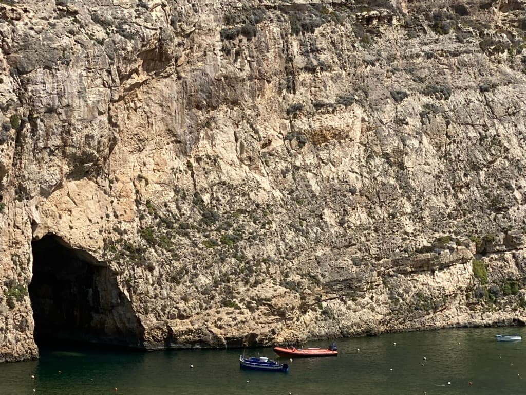 The Inland Sea lagoon — luzzu fishing boats moored in front of the tunnel entrance, the limestone cliff rising above the sheltered water