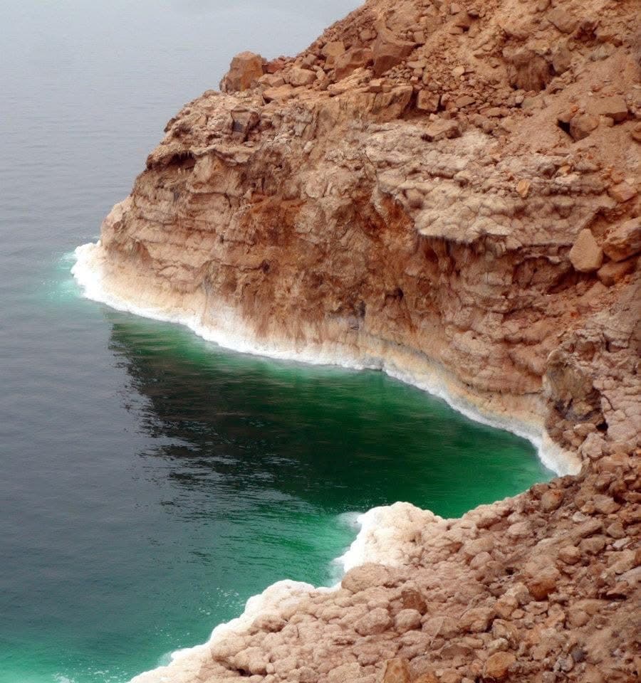 Salt-encrusted rock face dropping to the turquoise-green water of the Dead Sea — the colour a product of extreme salinity and mineral content