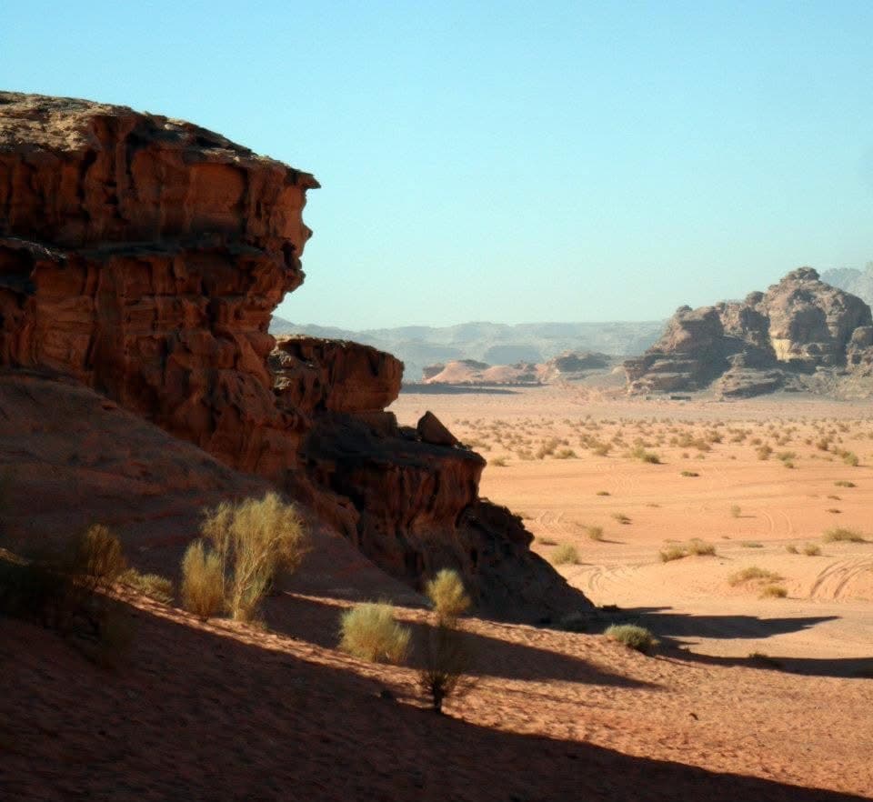 Red sandstone cliff face above the Wadi Rum desert floor — rock formations receding into the valley behind, the scale of the landscape consistently underestimated