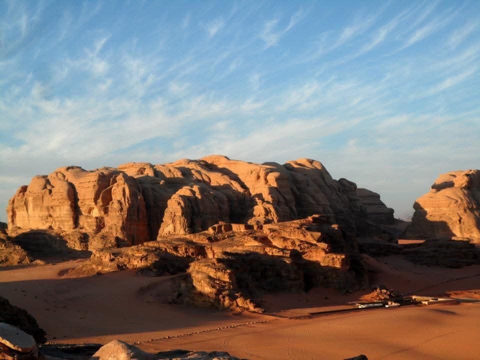 Wadi Rum at golden hour — layered sandstone formations rising from the desert floor, a Bedouin camp at the base of the rock
