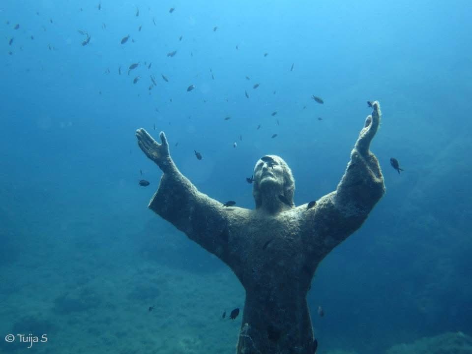 Il Cristo degli Abissi from the front — anemones on the hands, small wrasse working the folds of the robe, and a grouper stationed at the base of the plinth