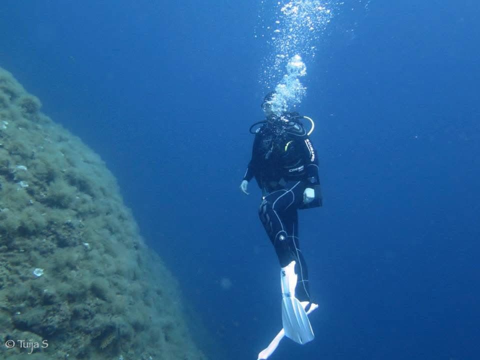 Diver ascending the vertical wall — bubbles rising into the blue-green Ligurian water, wall dropping beyond sport diving depth