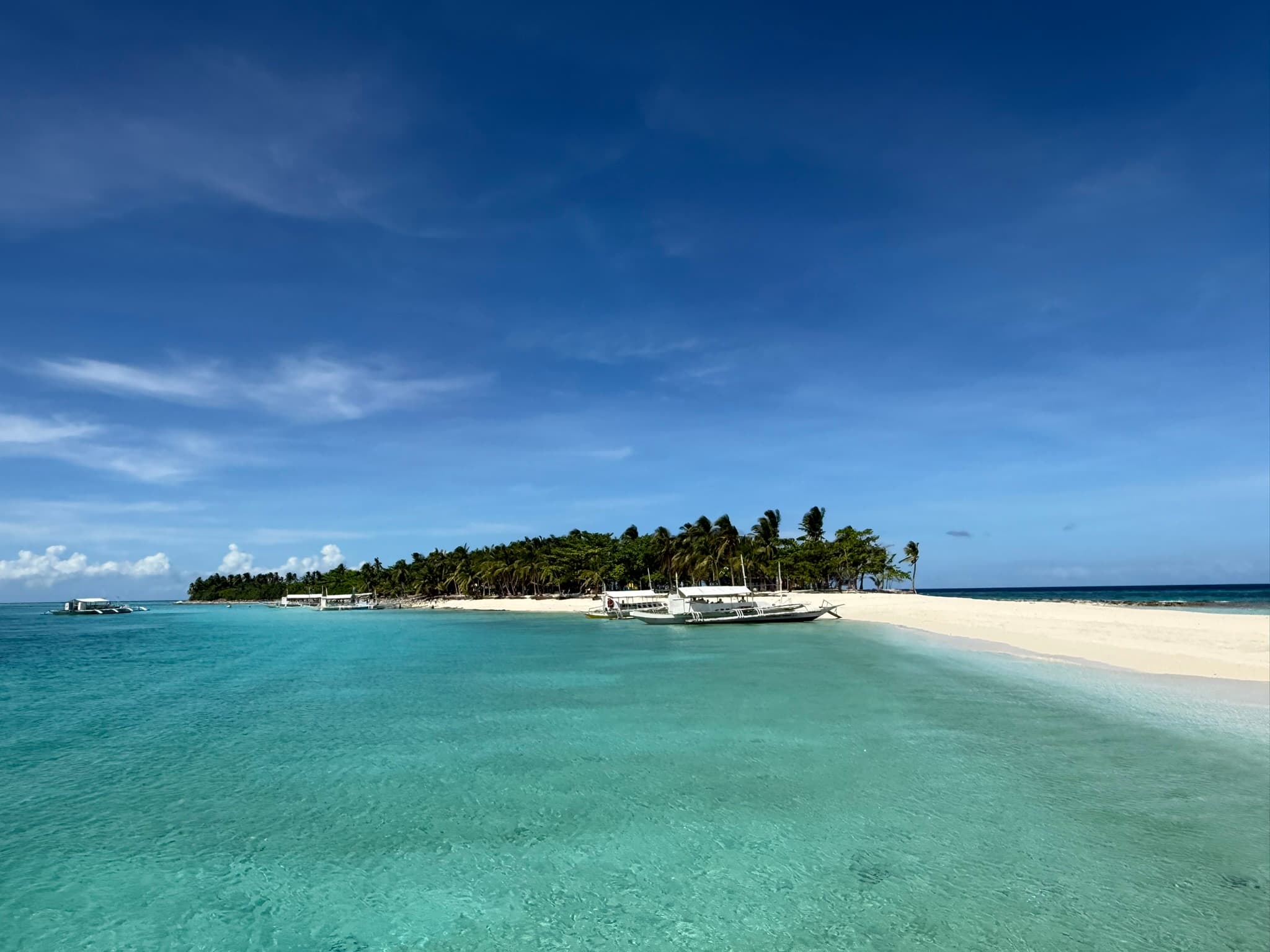 Kalanggaman Island from the water — bangka boats moored at the white sand shore, the palm-lined interior rising behind