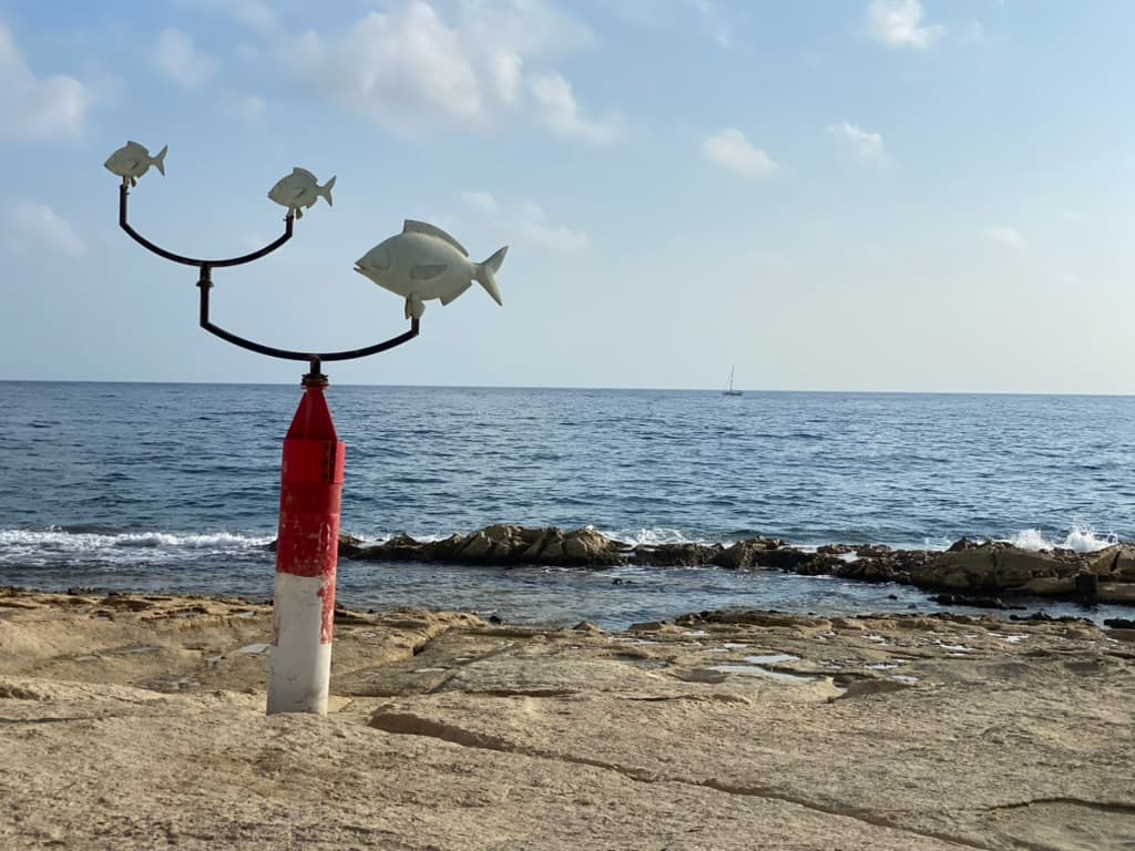 Dive site marker on the Maltese shoreline — a sculpture of three fish on a buoy post marking the shore entry point