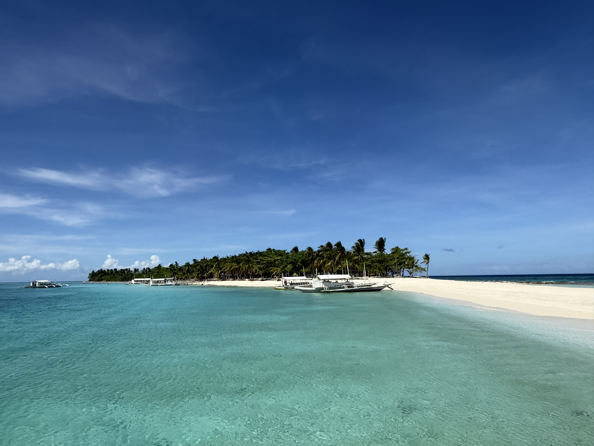 Kalanggaman Island from the water — bangka boats moored at the white sand shore, the palm-lined interior rising behind