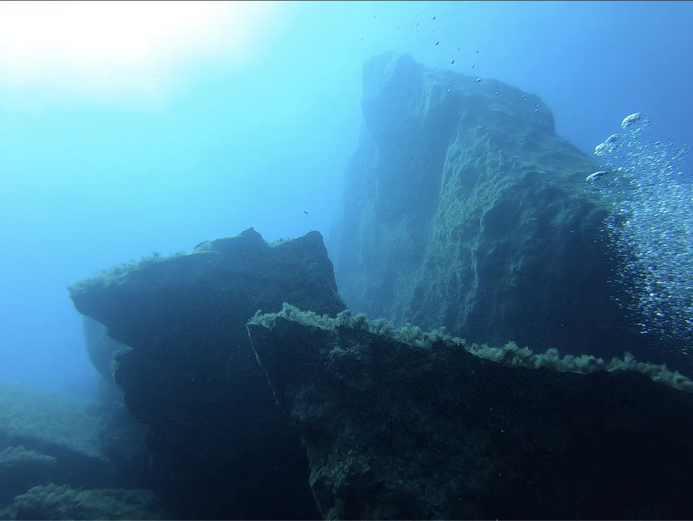 Wreck superstructure at depth — sponge growth on the upper hull, small fish schooling in the haze, sunlight filtering from the surface
